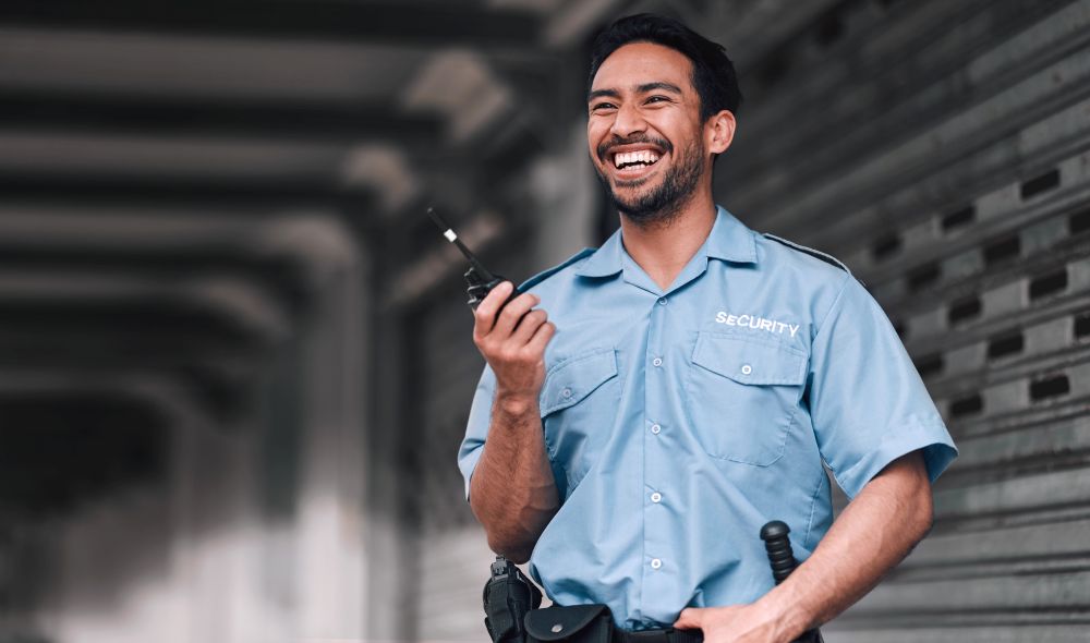 Smiling security guard in a light blue uniform holding a walkie-talkie stands against a background of a metal shutter.