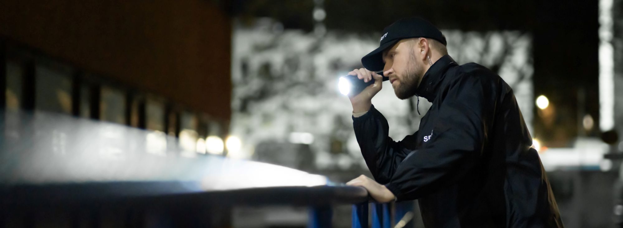 A person in a black jacket and cap shines a flashlight into the distance at night. They stand near a railing with blurred buildings and lights in the background.