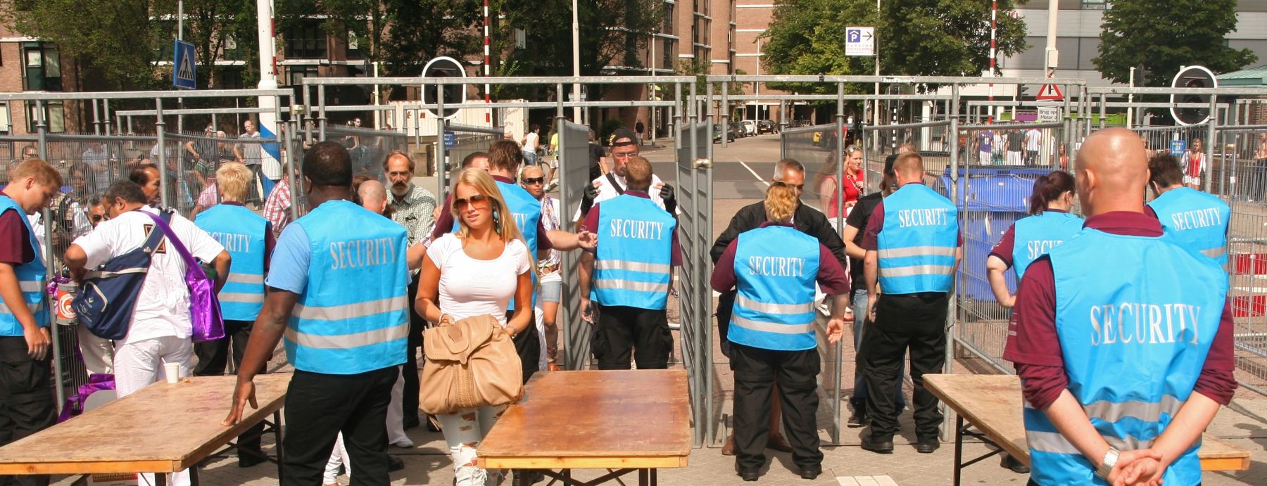 A group of security personnel in blue vests monitor people passing through a checkpoint with tables and barriers. Some people are carrying bags. Urban setting with trees and buildings in the background.