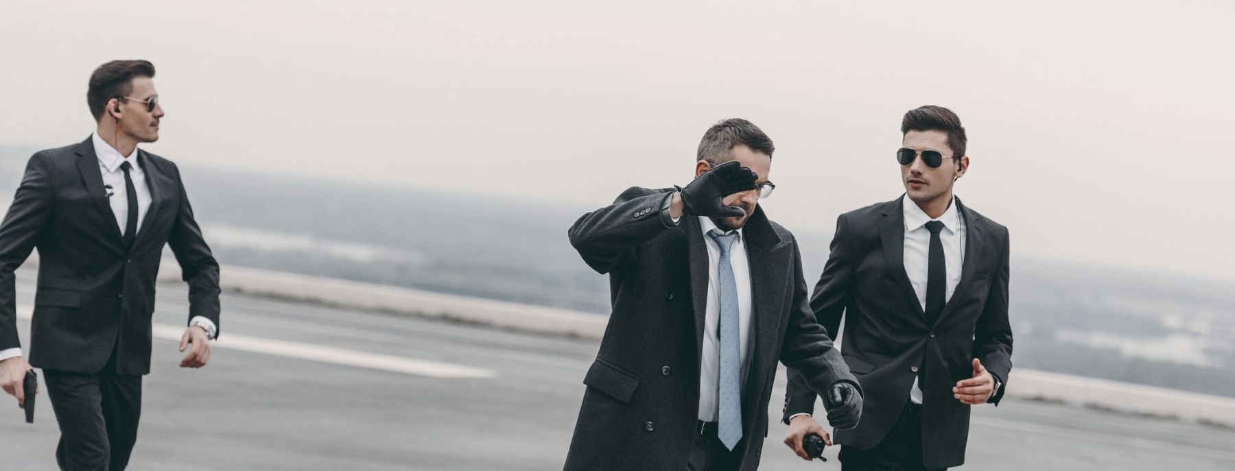 Three men in suits and sunglasses walk assertively on a rooftop. Two have earpieces, suggesting security personnel. The man in the middle gestures, partially covering his face with his hand. The sky is overcast, and the background is blurred.
