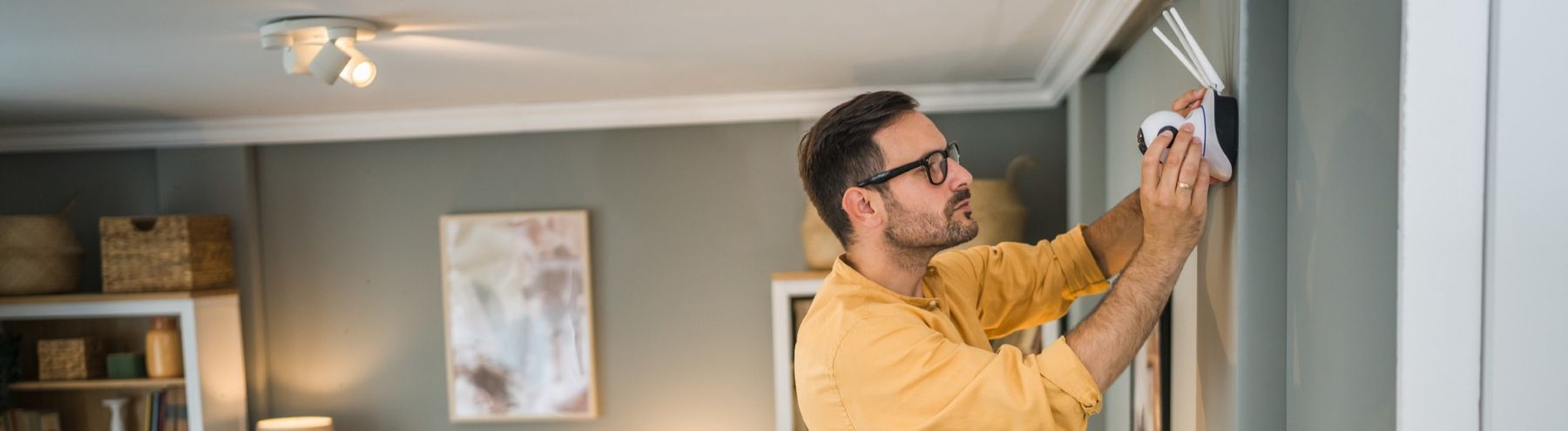 A man wearing glasses and a yellow shirt is installing a smoke detector on a gray wall in a living room. The room has modern decor, including shelves with baskets and a framed abstract painting.