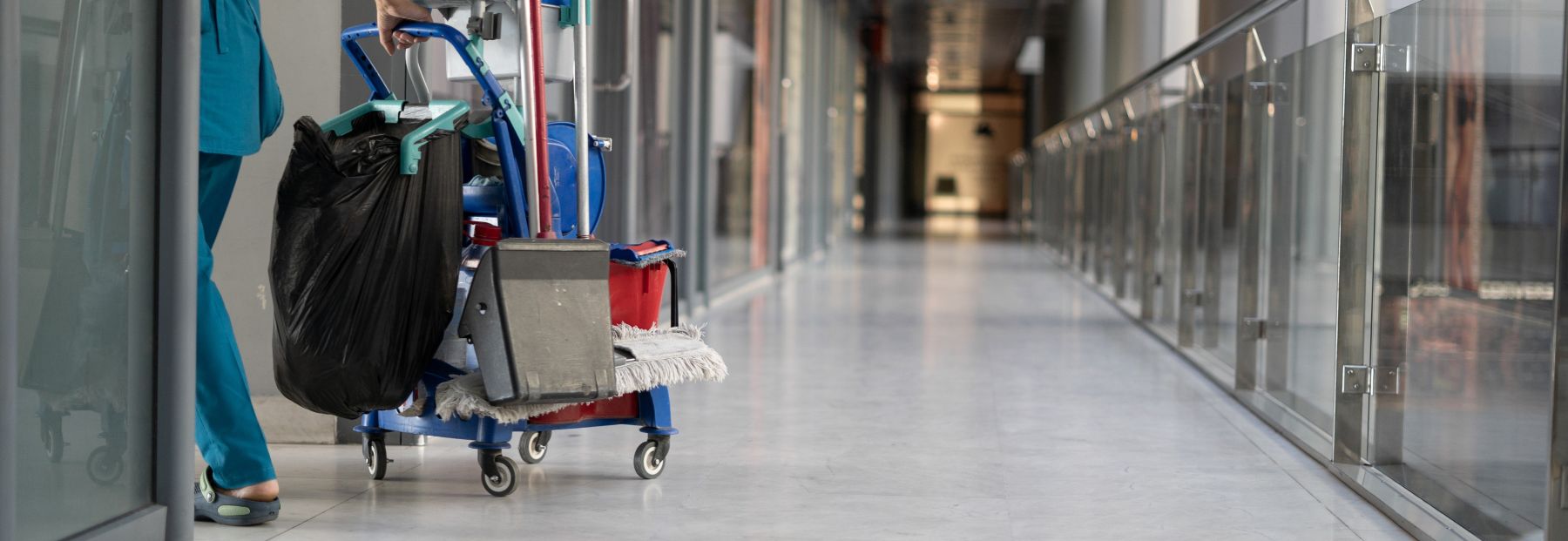 A janitor pushes a cleaning cart down a long, empty corridor with glass railings. The cart is equipped with various cleaning supplies, including mops, buckets, and a trash bag. The corridor is well-lit with a modern design.