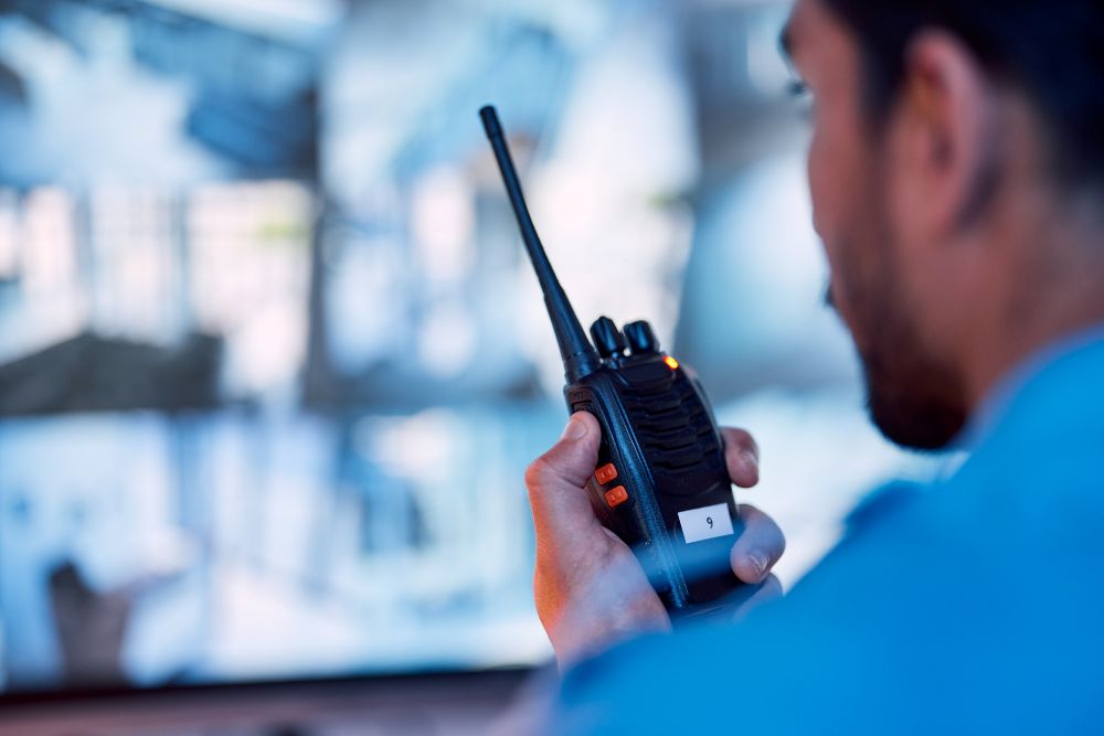 A person holding a walkie-talkie is looking at surveillance monitors in a control room. The image is focused on the walkie-talkie with blurred screens in the background displaying security footage.