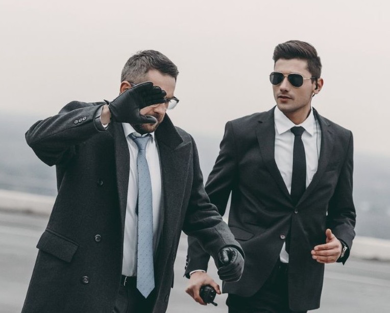 Two men in suits walking outdoors. One wears gloves and raises his hand to shield his face. The other wears sunglasses and an earpiece, holding a phone. The background is overcast.