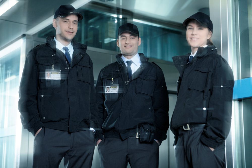 Three individuals in security uniforms stand together in an indoor setting. They wear black jackets, ties, and caps, each displaying an ID badge. The environment appears professional, with a slight motion blur in the image.
