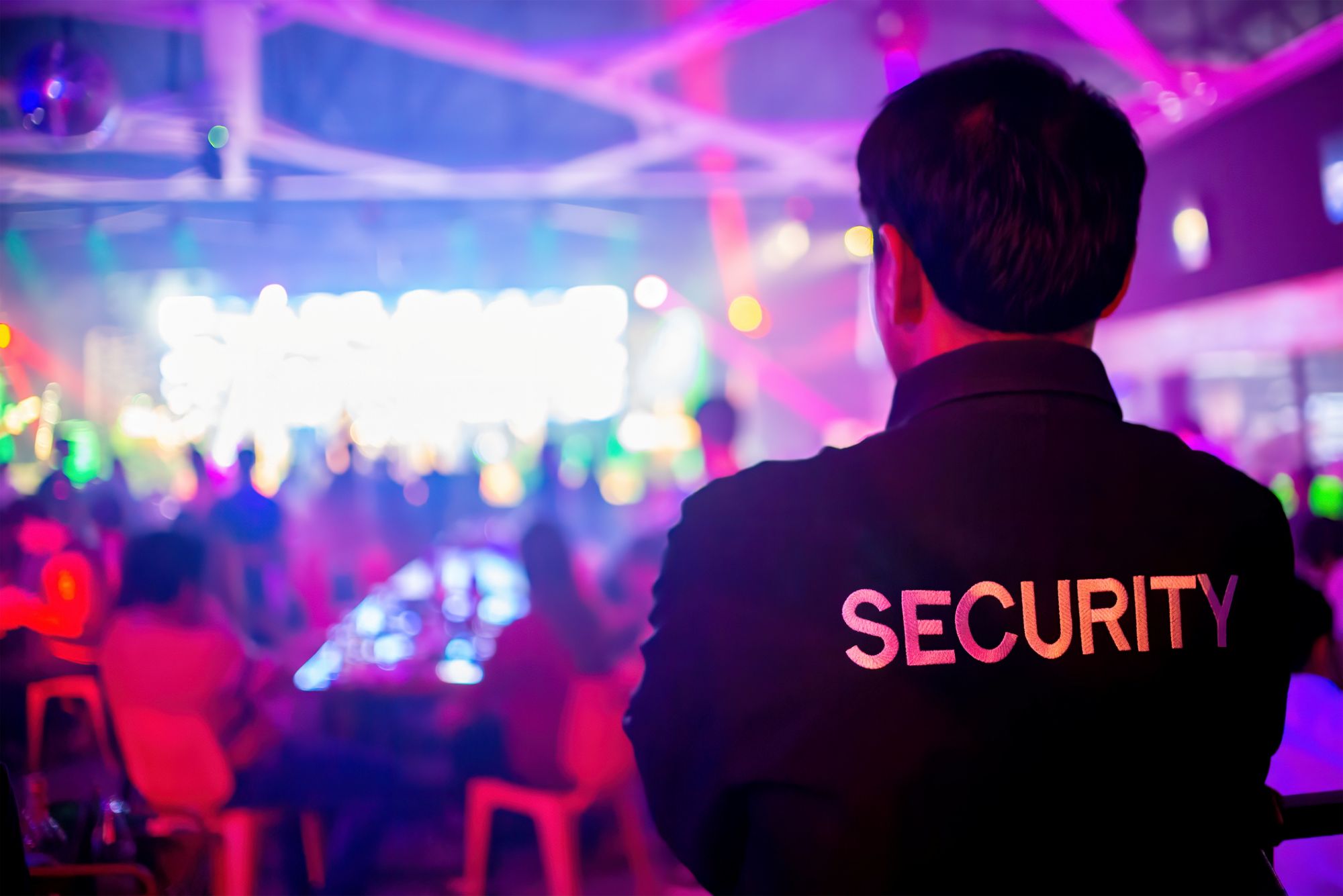 A security guard stands with "SECURITY" printed on the back of their shirt, overseeing a vibrant, colorful nightclub scene filled with people, lights, and movement.