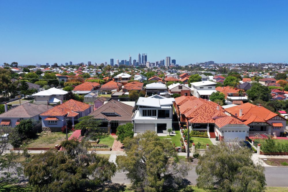 Aerial view of a suburban neighborhood with red and brown rooftops, lined with green trees. In the distance, a city skyline with tall buildings is visible under a clear blue sky.