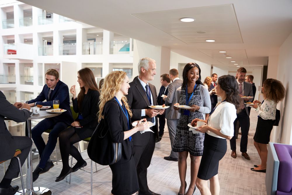 A group of professionally dressed people socialize in a modern office space. Some are standing with plates of food and drinks, engaged in conversation around high tables. The background features a bright, open area with multiple floors and walkways.