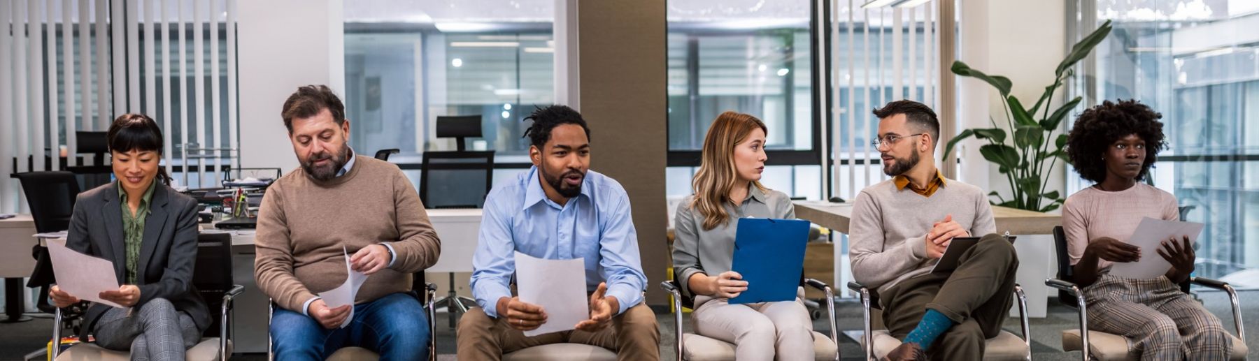 Six people are seated in an office waiting area, each holding papers. They appear to be engaged in conversation or waiting for an interview. The background shows office furniture and large windows.